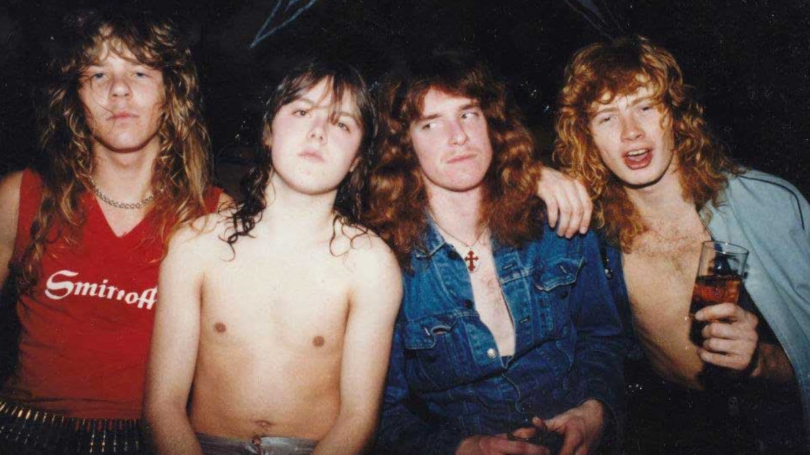 A 1983 photo of Metallica members. From left to right: James Hetfield with wavy shoulder-length hair in a red 'Smirnoff' sleeveless top and studded belt; Lars Ulrich, bare-chested with wavy dark hair; Cliff Burton with wavy red hair wearing a denim jacket with a cross necklace; and Dave Mustaine with curly blond hair, half in a blue shirt, holding a glass. Their relaxed poses and styles reflect the heavy metal culture of the time.